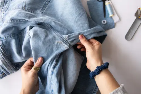 Hände reparieren ein Loch in Jeans mit einem Flicken. Nähutensilien liegen bereit.