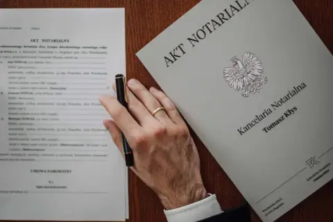 Akt poświadczenia dziedziczenia i umowa darowizny na biurku notariusza. Dłoń z obrączką trzyma długopis.