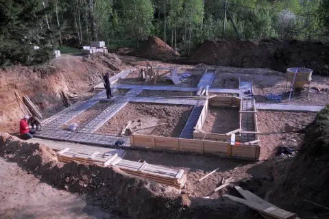 Budowa fundamentów domu. Widać zbrojenie i szalunki. Pracownicy przygotowują teren. Chudziak w tle.