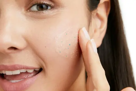 En kvinna applicerar en ansiktsskrubb med små, vita pormaskar-liknande korn för att exfoliera huden.