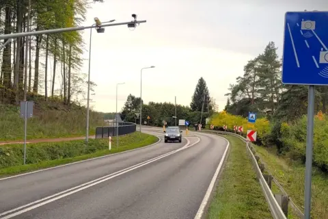 Znak informuje o systemie radarowym pomiaru prędkości. Na drodze widać samochód i kamery nad drogą.