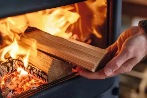 Eine Hand legt Holz in einen brennenden Kaminofen. Die Flammen lodern hell und wärmend.