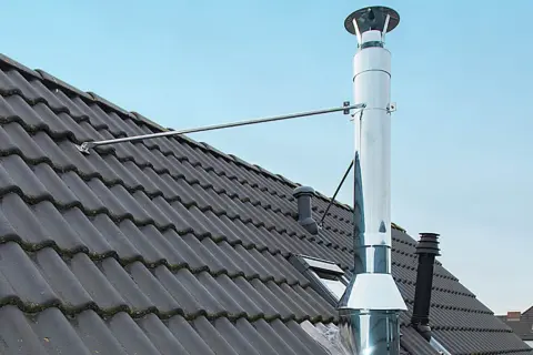 Edelstahl-Ofenrohre verbinden sich auf einem dunklen Ziegeldach. Ein Metallarm stützt das glänzende Rohr.