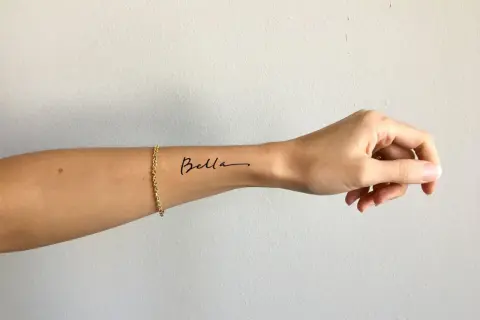 Ein zartes Tattoo-Armband mit dem Namen "Bella" auf dem Handgelenk, geschmückt mit einer goldenen Kette.