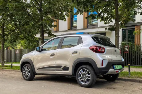 Srebrna Dacia Spring zaparkowana pod drzewami. Ten elektryczny samochód oferuje dobry zasięg na miejskie podróże.