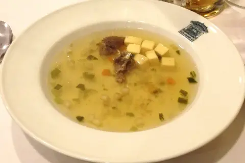 Un bol de potage clair avec des morceaux de viande, des dés de fromage, des légumes verts et des carottes.
