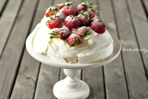 Pyszna beza pavlova z bitą śmietaną i świeżymi truskawkami, oprószona cukrem pudrem, prezentuje się apetycznie na białej paterze.