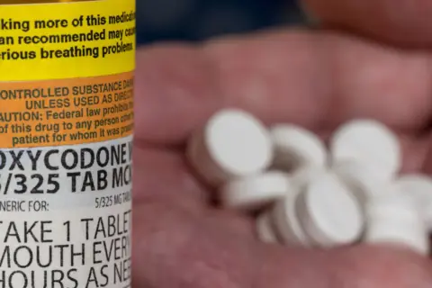 Butelka z lekiem Oxycodone 5/325 mg i garść białych tabletek w dłoni.