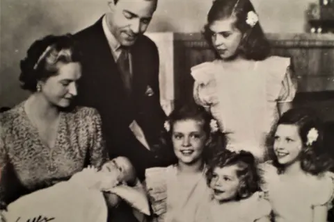 Familjefoto med prinsessan Sibylla och hennes barn. Bilden visar en familj med föräldrar och flera barn, inklusive en bebis. Prinsessan Sibylla bar en mönstrad klänning.