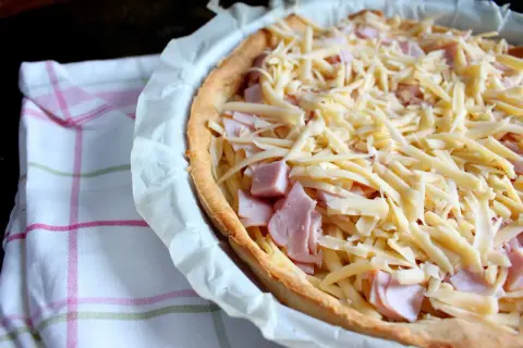 Tarta z szynką i serem, gotowa do pieczenia. Kruche ciasto, kawałki szynki i obfita warstwa startego sera.