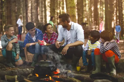 Kinder lachen und trinken am Lagerfeuer. Ein Lehrer verweigert die Klassenfahrt, aber die Kinder genießen den Ausflug trotzdem.