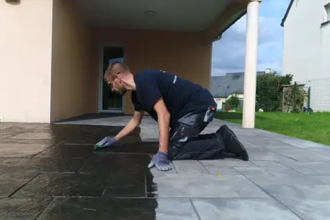 Un homme agenouillé effectue le nettoyage de pierre naturelle extérieur avec une éponge, redonnant son éclat à la terrasse.