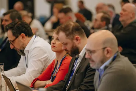 Ludzie siedzący przy stołach, słuchający prelekcji.