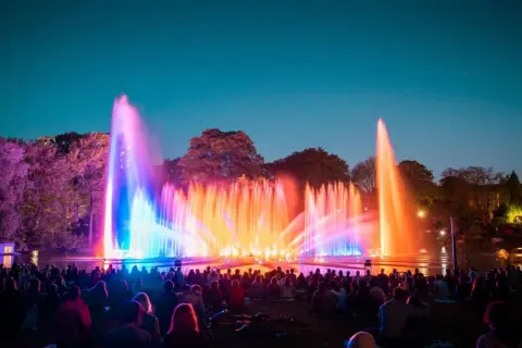 Spektakuläre Wasserspiele in Planten un Blomen erhellen den Abendhimmel in bunten Farben. Eine Menschenmenge sitzt im Gras und bestaunt die Lichtshow.