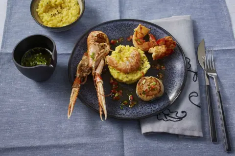 Ein Teller mit frutti di mare: Kaisergranat, Garnelen und Jakobsmuschel auf Safranrisotto, garniert mit Tomatenwürfeln. Daneben ein Schälchen mit Sauce und ein Schälchen Risotto.