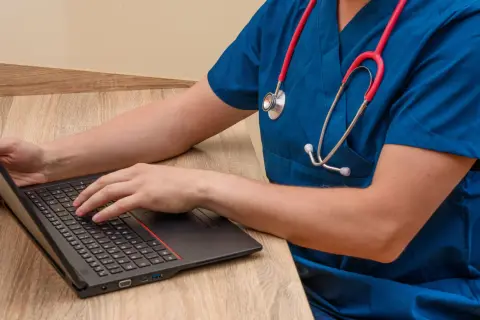 Lekarz w niebieskim fartuchu z czerwonym stetoskopem pisze na laptopie, szukając informacji, jaki lekarz zarabia najwięcej.