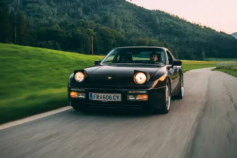 Dunkelroter Porsche mit Klappscheinwerfer fährt auf kurviger Landstraße durch grüne Hügel.