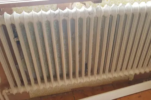 Gammal vit element med blommig tapet bakom. Kanske dags att ta bort elementet och tapeten för en renovering.