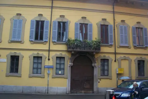Facciata gialla del **Palazzo Belgioioso** con finestre dalle persiane azzurre e un balcone fiorito.