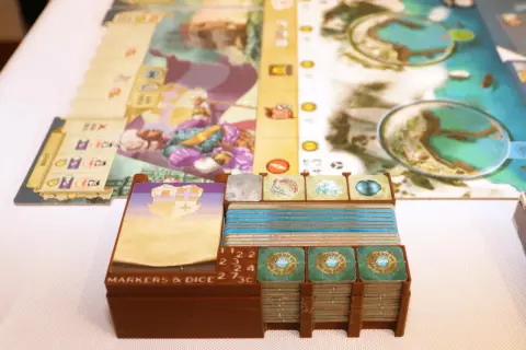 Spielkomponenten für das Dead Reckoning Brettspiel, darunter Karten, Marker und Würfel, sind ordentlich in braunen Halterungen gestapelt.