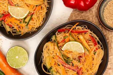 Zwei Schalen mit erfrischendem Glasnudel Salat, garniert mit Limettenspalten, Karotten- und Gurkenstreifen.
