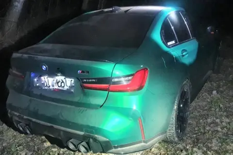 Zielony BMW M3 Competition, z napisem "bogus m3" na tylnej szybie, stoi nocą na leśnej drodze.