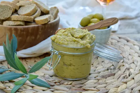 Un pot de tapenade verte, garni d'une cuillère en bois, est prêt à être dégusté avec des toasts et des olives.
