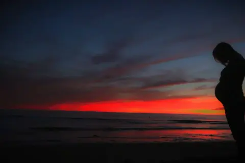 Kobieta w ciąży na tle zachodu słońca nad oceanem. Pamiętaj o ubezpieczeniu turystycznym w ciąży, by podróżować bezpiecznie.
