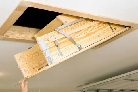 A wooden attic ladder is being installed, with a hand pulling down the first section.