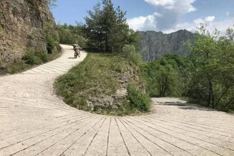 Ciclista su strada sterrata nel Parco Alto Garda Bresciano, tra rocce e vegetazione lussureggiante, con montagne sullo sfondo.