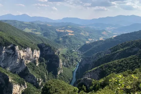 Vista mozzafiato dalla terrazza del Furlo: un fiume serpeggia tra gole rocciose e colline verdi, sotto un cielo azzurro con nuvole bianche.