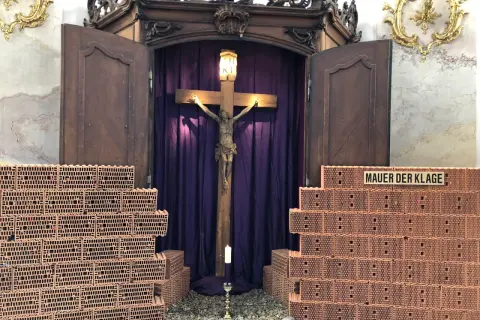 Ein Kreuz mit Jesusfigur, umgeben von Ziegelmauern und lila Vorhängen. Ein Kerzenlicht lädt zum Kreuz Gebet ein.
