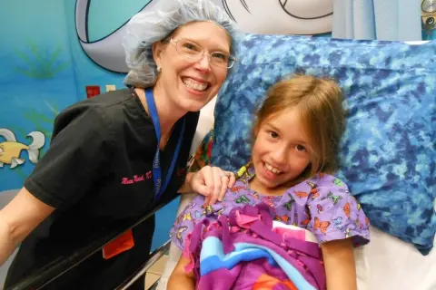Niña sonriente en cama de hospital, junto a enfermera. Ella tiene síndrome de Marfan y recibe cuidados.