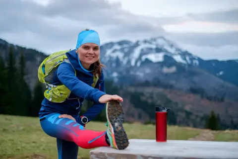 Uśmiechnięta biegaczka przygotowuje się do treningu ultramaraton 50 km, rozciągając nogę na tle gór.