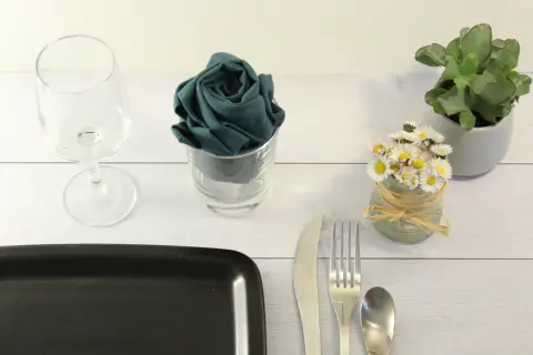 Un verre à pied, une serviette pliée en rose dans un verre, des petites fleurs et des couverts sur une table en bois.