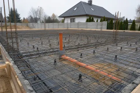 Zbrojenie i rury kanalizacyjne na poziomie zero budynku. W tle dom i ogrodzenie.