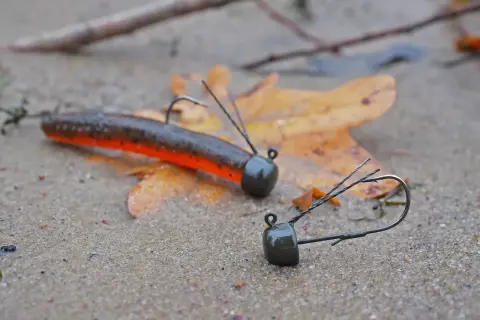 Zwei Angelköder, ein ned rig und ein weiterer, liegen auf nassem Sand neben einem Herbstblatt.