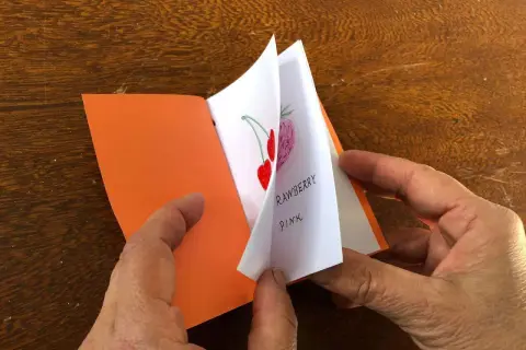 Mani che sfogliano un piccolo libro fatto a mano, con disegni di fragole e ciliegie. Un'idea per imparare come fare un libro a mano.