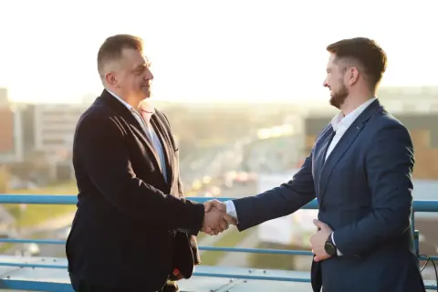 Dwóch mężczyzn w garniturach ściska sobie dłonie na tle miasta.