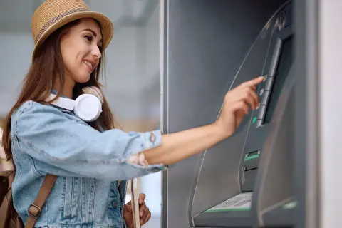 Uśmiechnięta kobieta w kapeluszu i dżinsowej kurtce dokonuje wypłaty z bankomatu za granicą.