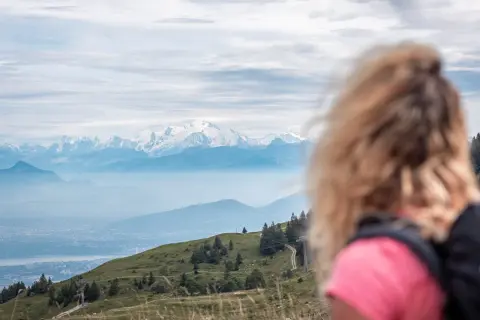 Vue panoramique sur les Alpes enneigées, avec une femme blonde en premier plan. Un sentier serpente sur la colline verdoyante, invitant au trek dans le Jura.