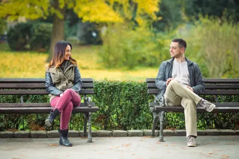 En kvinna och en man sitter på varsin parkbänk och ler mot varandra. Kanske funderar hon på hur blir man attraktiv som tjej, och han på hur blir man attraktiv som kille.