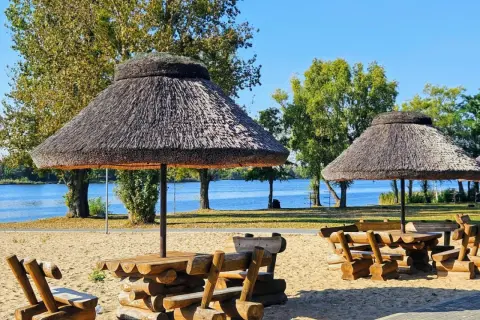 Drewniane stoły z ławami pod strzechą na plaży w Rogoźno Wielkopolskie, nad błękitną wodą.