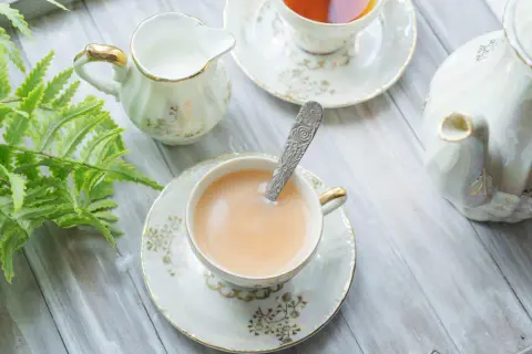 Elegancka filiżanka z herbatą z mlekiem, ozdobiona złotymi wzorami, obok dzbanuszka i czajniczka.
