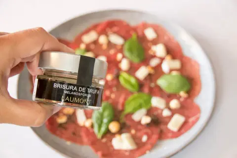 Mano sostiene un frasco de "Brisura de Trufa" Laumont, listo para añadir a un carpaccio de ternera con queso y albahaca.