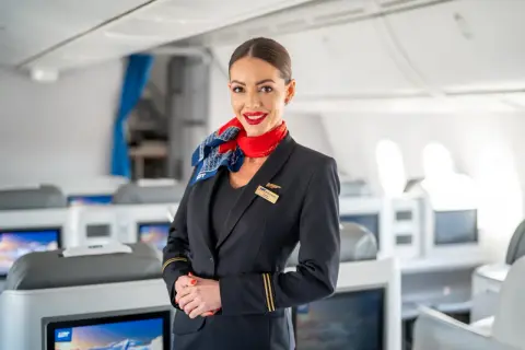 Uśmiechnięta członkini **cabin crew** w granatowym mundurze z czerwonym szalem, stoi na tle rzędów foteli w samolocie.