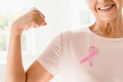 Lächelnde Frau mit rosa Schleife zeigt Bizeps. Onkologische Physiotherapie stärkt den Körper und gibt Hoffnung.