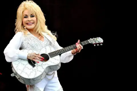 Dolly Parton, w białym, błyszczącym stroju, gra na gitarze. Wyobraź sobie, że śpiewa "Jolene", a tekst piosenki rozbrzmiewa w powietrzu.