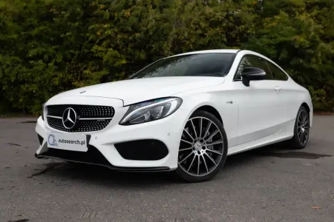 Biały Mercedes C43 AMG coupe z czarnymi felgami, zaparkowany na tle zieleni.