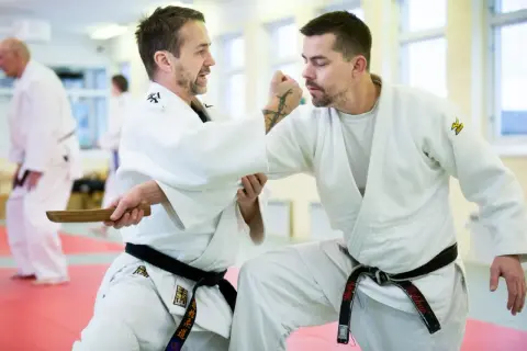 Två män i judodräkter tränar en judo kast. En av dem håller i ett träföremål.